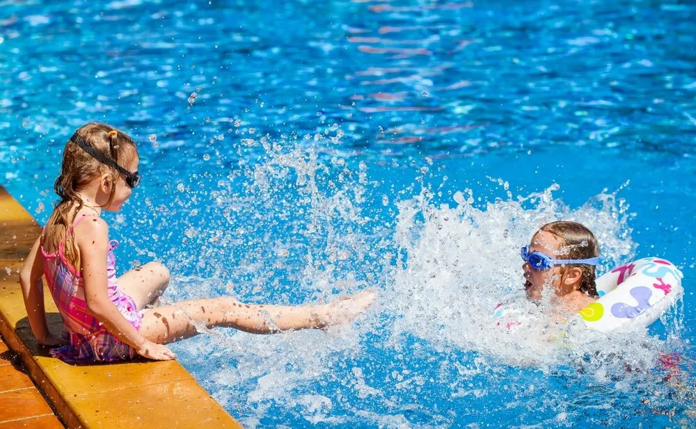 Two girls wearing goggles splash and play in a bright outdoor swimming pool.
